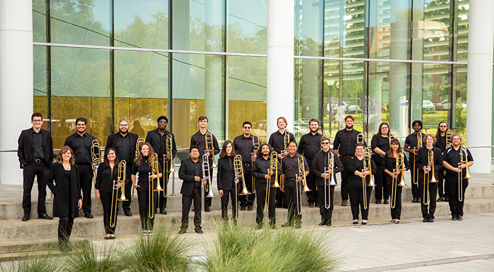 A large trombone ensemble standing in front of a glass wall